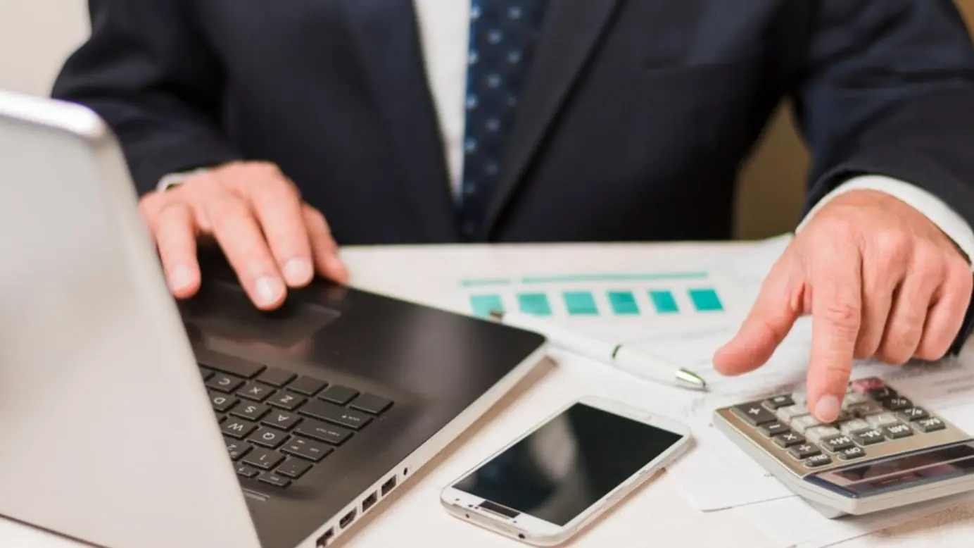 Un uomo usa una calcolatrice mentre lavora su un computer portatile. 